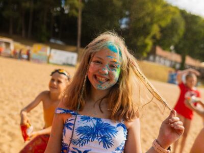 Uśmiechnięta dziewczynka z kolorowym proszkiem na twarzy na plaży. Radosne, wakacyjne chwile.