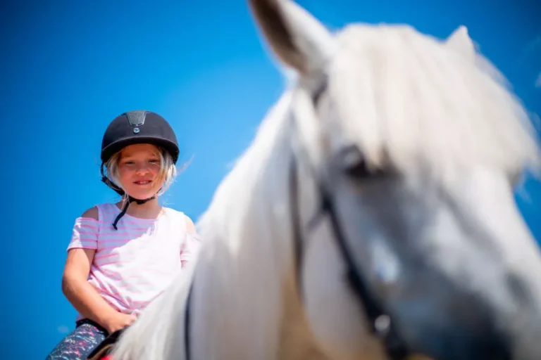 Dziewczynka w kasku na białym koniu pod błękitnym niebem. Jazda konna, dzieci.