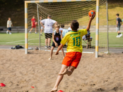 Chłopiec w żółtej koszulce z numerem 10 rzuca piłką do ręcznej na piasku.