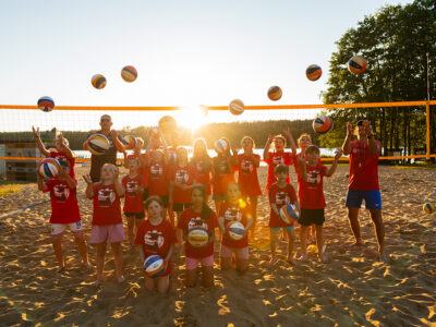 Dzieci i trener bawią się piłkami do siatkówki na plaży w słoneczny dzień.