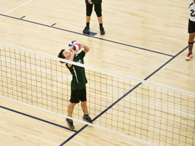 Chłopiec w zielonej koszulce serwuje piłkę do siatkówki na drewnianej hali sportowej.