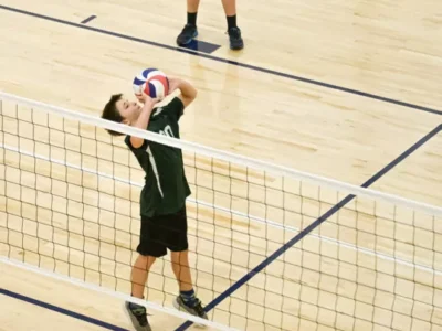 Chłopiec w zielonej koszulce serwuje piłkę do siatkówki nad siatką.