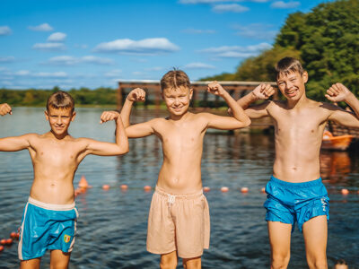 Trzech chłopców pozuje nad jeziorem, pokazując siłę. Letnie wakacje, zabawa nad wodą.