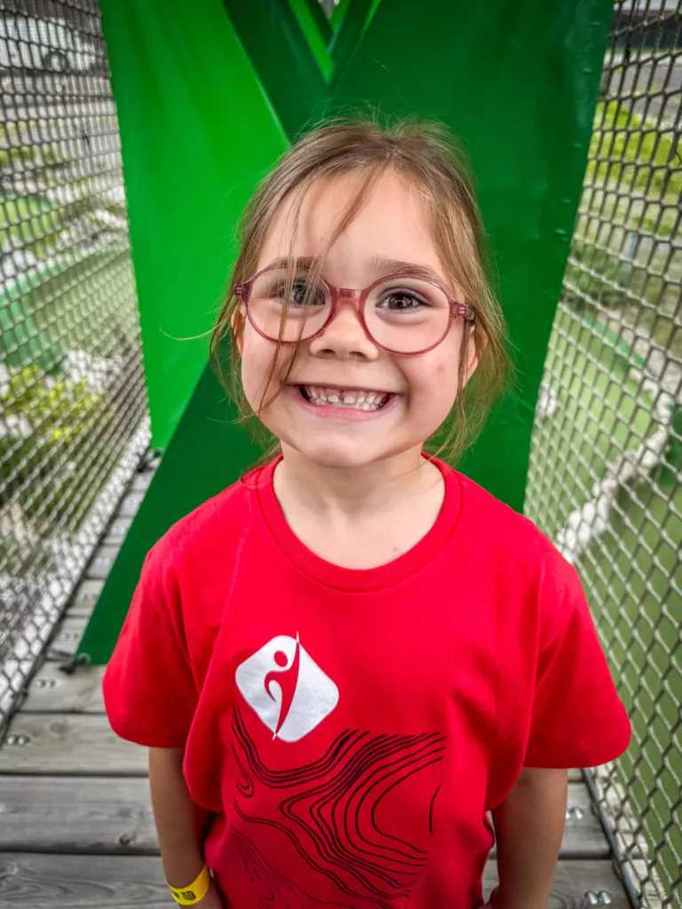Uśmiechnięta dziewczynka w okularach i czerwonej koszulce na tle zielonej konstrukcji.
