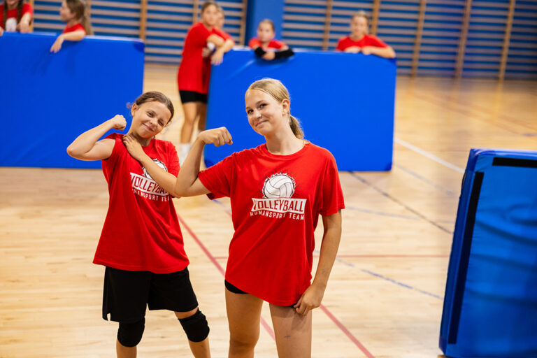 Dwie dziewczynki w czerwonych koszulkach z logo siatkówki pokazują siłę na sali gimnastycznej.