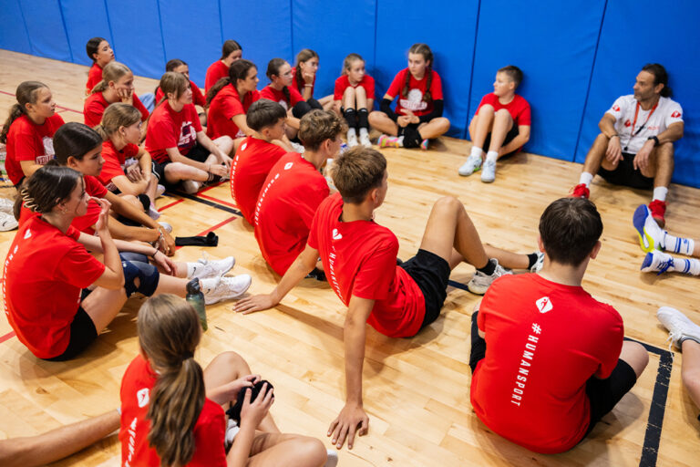 Dzieci w czerwonych koszulkach (#HUMANSPORT) siedzą na podłodze, słuchając trenera.