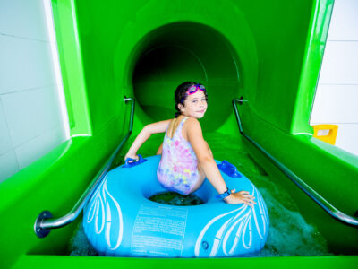 Dziewczynka z kółkiem zjeżdża po zielonej zjeżdżalni wodnej.