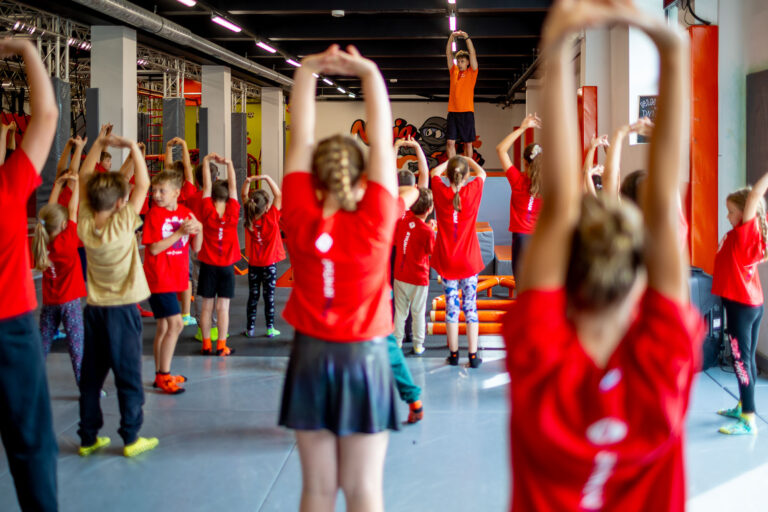 Dzieci w czerwonych koszulkach ćwiczą z trenerem w sali gimnastycznej.