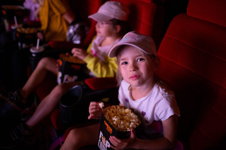 Dziecko w kinie z popcornem. Uśmiechnięta dziewczynka w czapce.