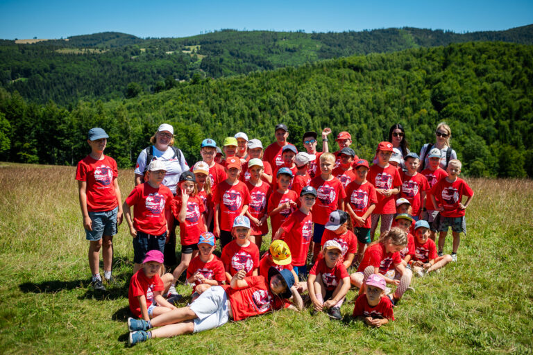 Grupa dzieci w czerwonych koszulkach na tle zielonych wzgórz.