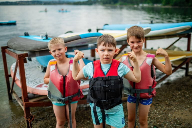Trzech chłopców w kamizelkach ratunkowych pozuje z uśmiechem nad jeziorem, gotowi na wodne przygody.