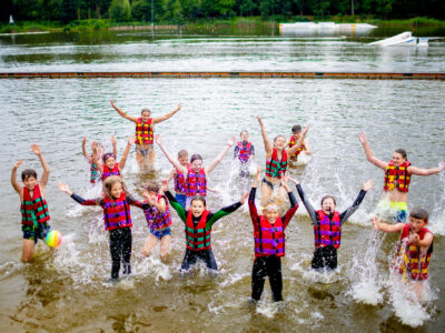 Dzieci w kamizelkach ratunkowych pluskają się w wodzie, radośnie unosząc ręce.
