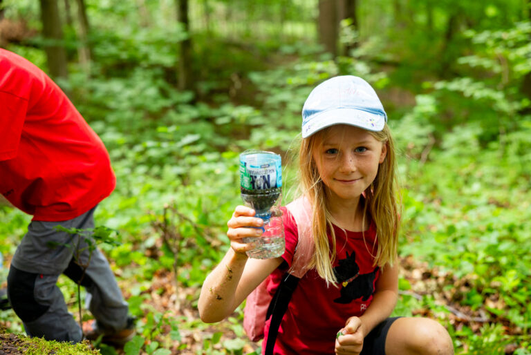 Dziewczynka w czapce z daszkiem trzyma butelkę wody w lesie.