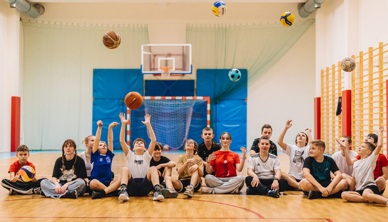 Dzieci na sali gimnastycznej rzucają piłki do góry, radość i sport.