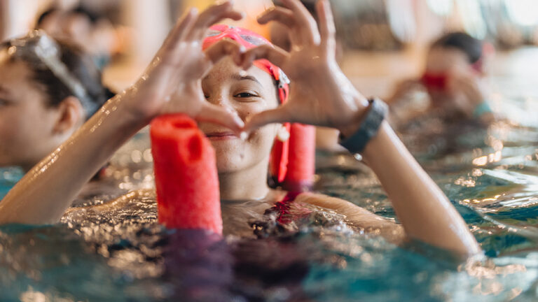 Dziewczynka w basenie tworzy serce dłońmi. Radość z nauki pływania.