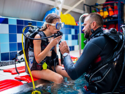 Nauka nurkowania: instruktor pomaga dziecku założyć sprzęt do nurkowania w basenie.