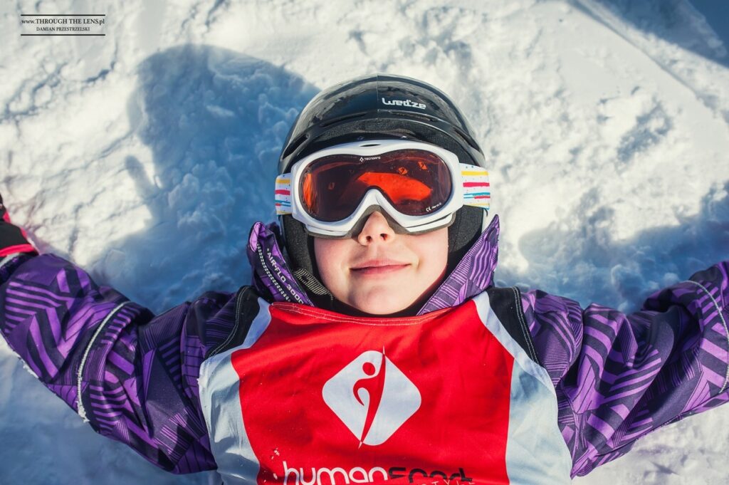 Dziecko w goglach narciarskich i kasku leży na śniegu.