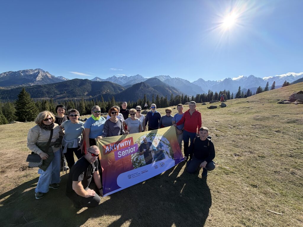 Grupa seniorów z banerem "Aktywny Senior" na tle górskiego krajobrazu.